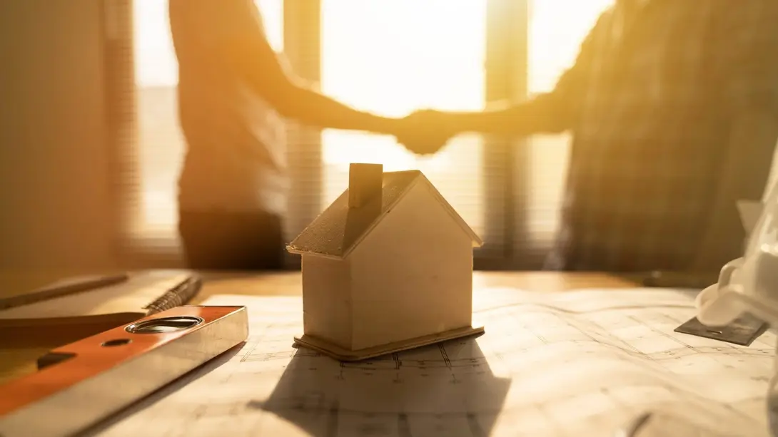Hausbau-Beratung mit Handschlag Hausmodell auf Bauplänen mit Handschlag im Hintergrund bei einem Beratungsgespräch.