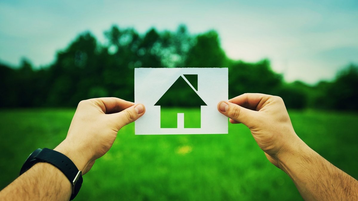 Hände halten ein Papiersymbol eines Hauses vor einer grünen Wiese.