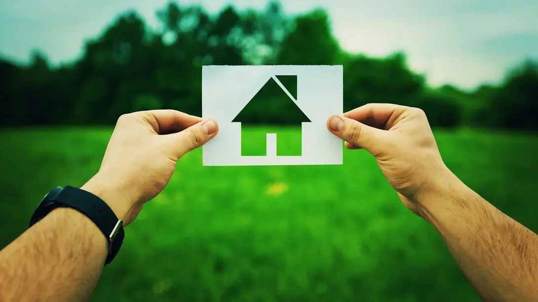 Hände halten ein Haus-Symbol vor einer grünen Wiese