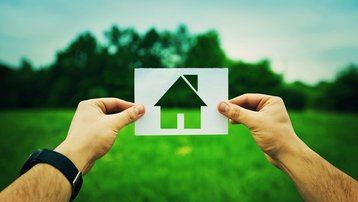 Hände halten ein Papiersymbol eines Hauses vor einer grünen Wiese.