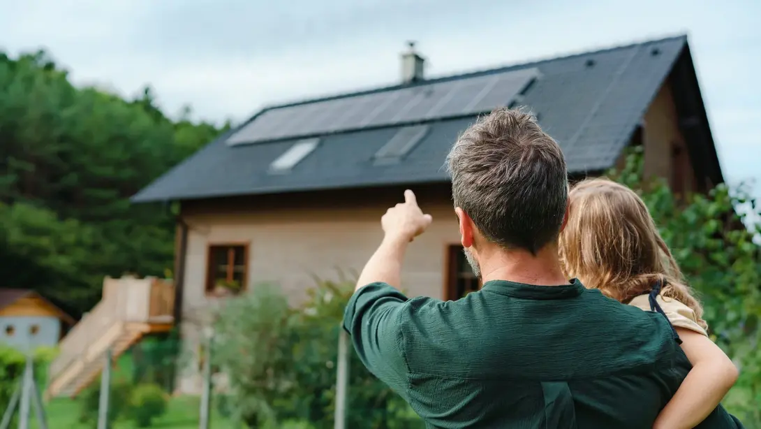 Nachhaltiges Eigenheim mit Photovoltaik Vater mit Kind blickt auf ein Haus mit Photovoltaikanlage in naturnaher Umgebung.