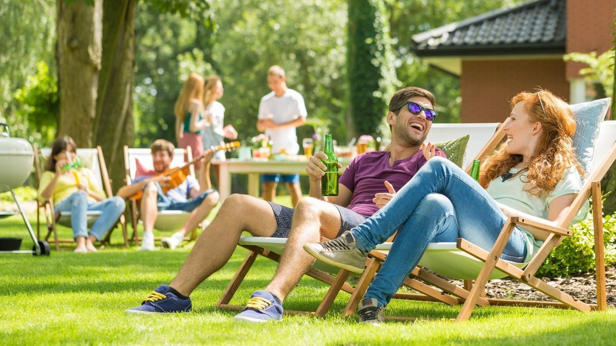 Freunde entspannen im Garten auf Liegestühlen mit Grill und Sitzbereich im Hintergrund