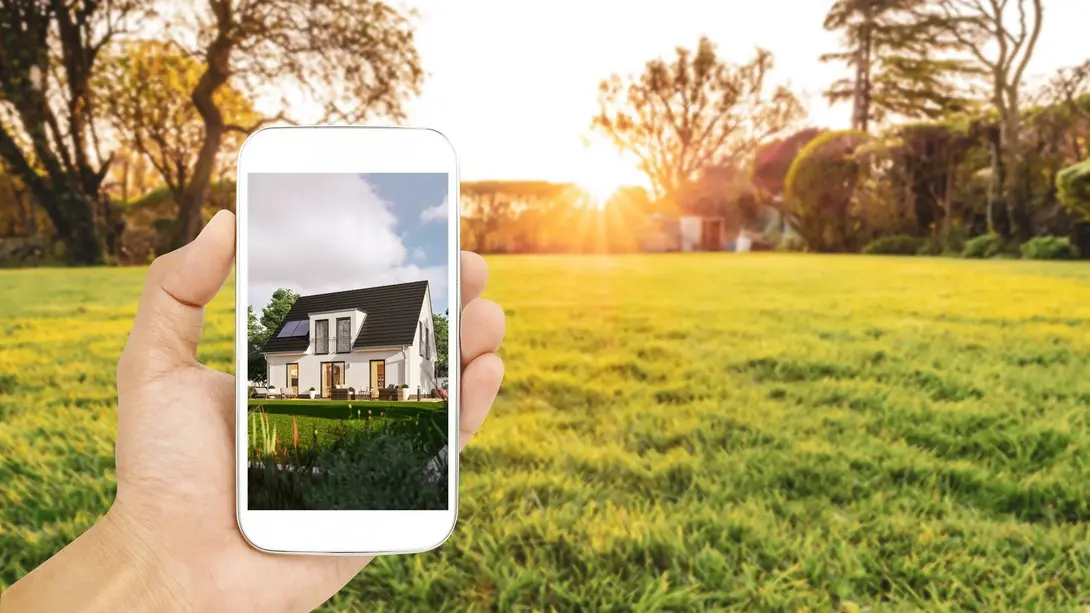 Hausplanung auf dem Grundstück visualisieren Hand hält ein Smartphone mit Hausentwurf vor einer freien Wiese bei Sonnenuntergang.