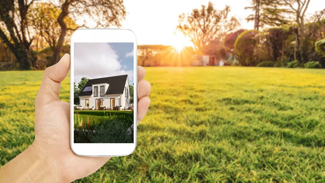 Hausplanung auf dem Grundstück visualisieren Smartphone mit Hausvisualisierung vor einer grünen Wiese bei Sonnenuntergang