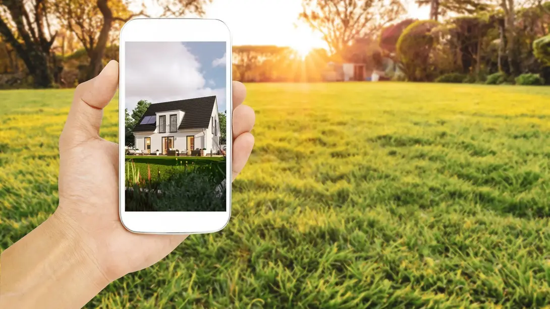 Hand hält ein Smartphone mit Hausentwurf vor einer freien Wiese bei Sonnenuntergang.