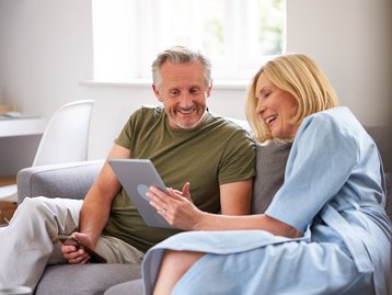 Älteres Paar sitzt entspannt auf dem Sofa und betrachtet gemeinsam ein Tablet.