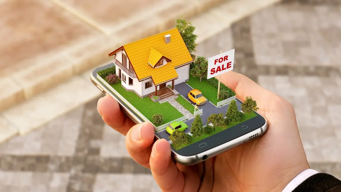 Digitale Suche nach Haus und Grundstück Hand hält ein Smartphone mit Hausmodell und Verkaufsschild als Symbol für digitale Grundstücks- und Immobiliensuche.