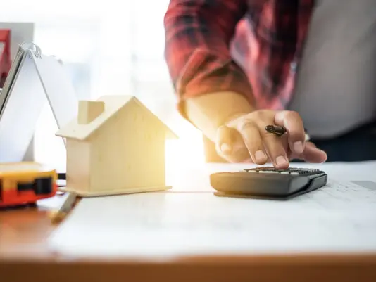 Person berechnet die Kosten für den Hausbau neben einem Hausmodell und Bauplänen
