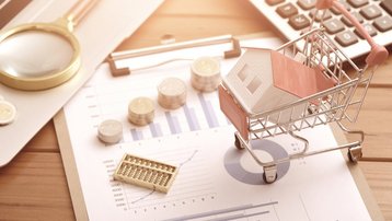 Hausmodell im Einkaufswagen neben Rechner, Münzen und Finanzunterlagen zur Baufinanzierung